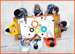 Various people sitting around a table working together on a white page with multicolored gears on it.