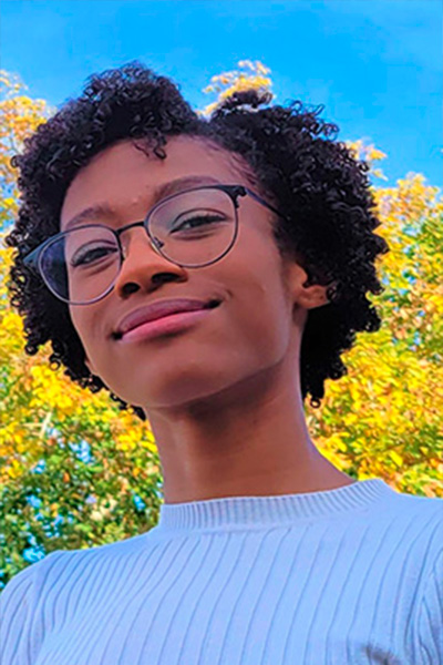 Kayla Richardson, a senior at Arizona State University who is poised to graduate with a degree in Electrical Engineering with a focus on Power and Energy. She has brown skin, short, curly hair, and glasses. She is pictured smiling, outdoors.
