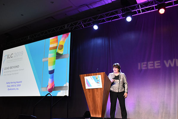 A speaker stands at a podium in front of  a purple curtain with the IEEE WIE logo on it at the ILC 2019 conference.