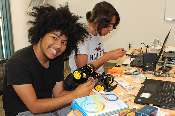 Two students work on electronics at a table. One is smiling and looks at the camera.