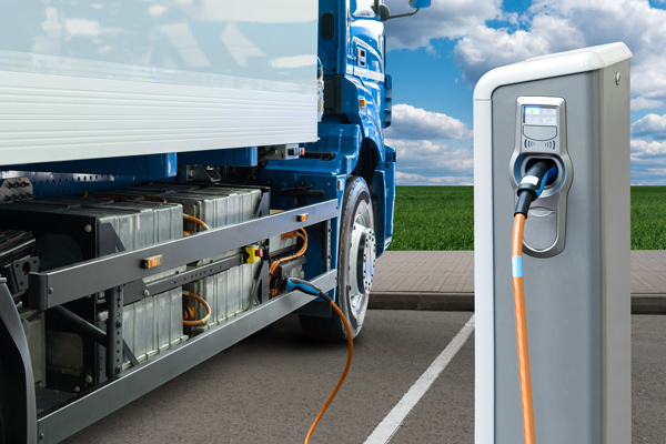 A truck is plugged into an electric charging station, with a blue sky with white clouds in the background.