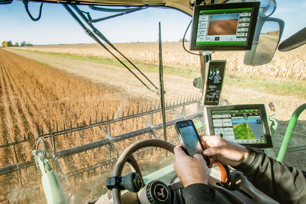 Farmer’s hands controlling a smart phone from a farming vehicle. Other screens showing farming process status in frame. 